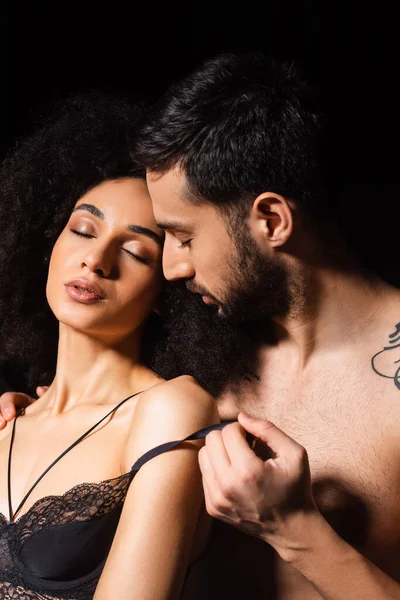 Bearded man undressing sensual african american girlfriend isolated on black — Stock Photo