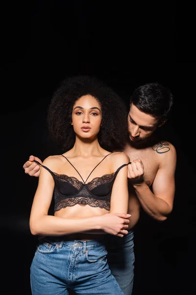 Hombre apasionado quitándose el sujetador de la mujer afroamericana aislado en negro - foto de stock
