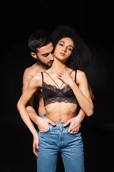 Homem barbudo beijando pescoço de mulher americana africana em jeans e sutiã isolado em preto — Fotografia de Stock