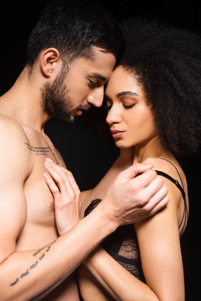 Hombre sin camisa quitándose el sujetador de la mujer afroamericana aislado en negro - foto de stock