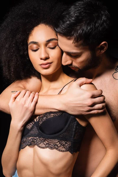 Hombre sin camisa abrazando seductora afroamericana mujer aislada en negro - foto de stock
