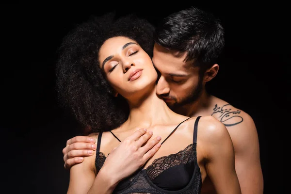 Hombre tatuado besando cuello de hermosa novia afroamericana en sujetador aislado en negro - foto de stock