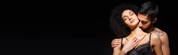 Young man kissing neck of african american woman in lace bra isolated on black, banner — Stock Photo