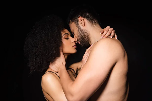 Vista lateral del hombre sin camisa besando mujer afroamericana aislada en negro - foto de stock