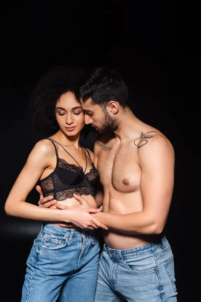 Bare chested man hugging seductive african american girlfriend on black background — Stock Photo