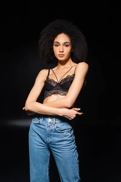 Sensual african american woman in jeans and bra looking at camera on black background — Stock Photo