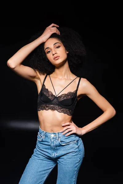 Giovane donna afroamericana in reggiseno e jeans guardando la fotocamera su sfondo nero — Foto stock