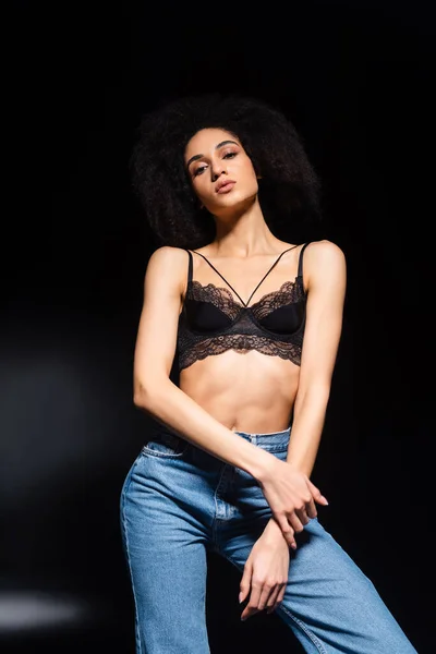 Low angle view of seductive african american woman in jeans and bra looking at camera on black background — Stock Photo