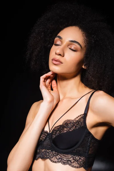 Mujer afroamericana en sujetador posando con los ojos cerrados sobre fondo negro - foto de stock