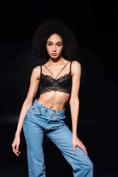 Sensuelle femme afro-américaine en jeans et soutien-gorge regardant la caméra isolée sur noir — Photo de stock