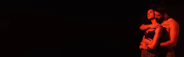 Uomo abbracciando ragazza afro-americana in reggiseno a luce rossa isolato su nero, banner — Foto stock