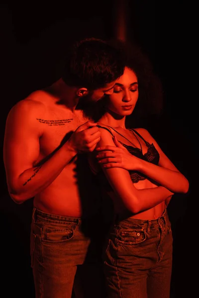 Muscular man taking off bra from sensual african american woman in red light on black background — Stock Photo