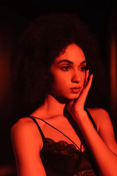 Femme afro-américaine en soutien-gorge en dentelle toucher le visage en lumière rouge sur fond noir — Photo de stock