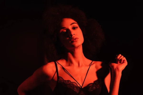 Pretty african american woman in lace bra looking at camera in red light isolated on black — Stock Photo