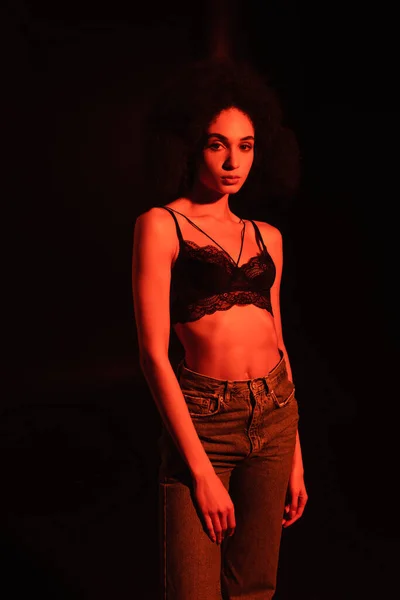 Young african american woman in jeans and bra looking at camera isolated on black with red lighting — Stock Photo