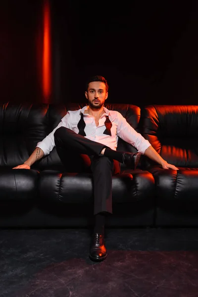 Бородатий чоловік у формальному одязі дивиться на камеру на дивані на чорному тлі з червоним освітленням — стокове фото