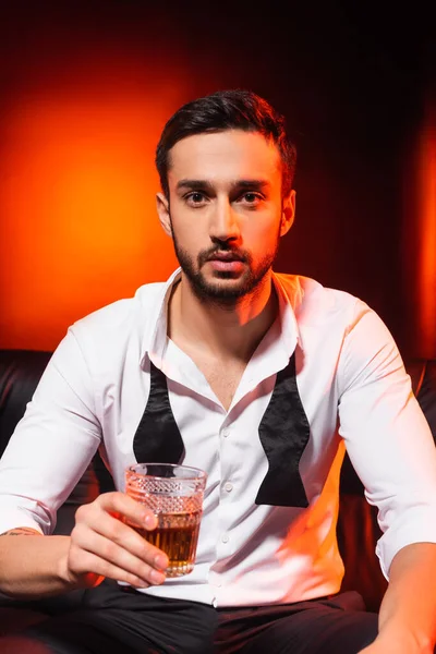 Uomo in abito formale che tiene un bicchiere di whisky sul divano su sfondo nero con illuminazione rossa — Foto stock