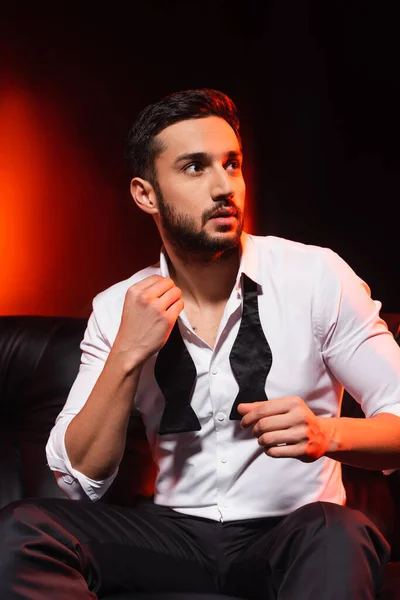 Elegant man adjusting bow tie on black background with red lighting — Stock Photo