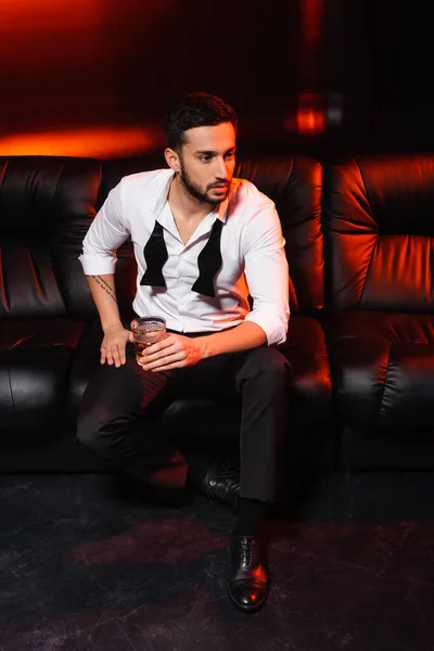 Man in formal wear holding glass of whiskey on couch on black background with red lighting — Stock Photo