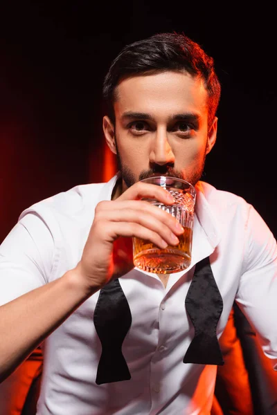 Hombre barbudo en ropa formal bebiendo whisky sobre fondo negro con iluminación roja — Stock Photo