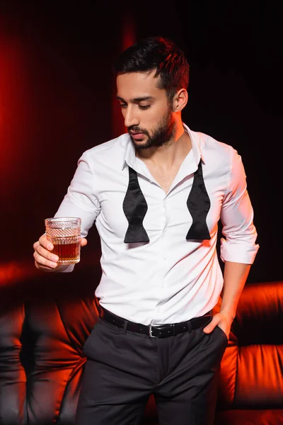 Elegante uomo che tiene un bicchiere di whisky vicino al divano su sfondo nero con illuminazione rossa — Foto stock