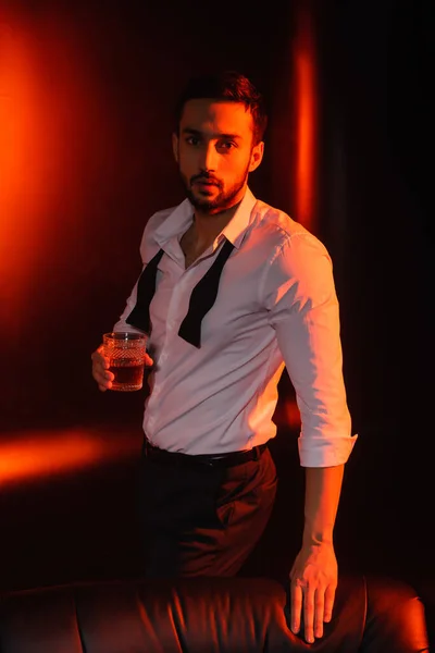 Uomo barbuto in camicia che tiene un bicchiere di whisky vicino al divano su sfondo nero con illuminazione rossa — Foto stock