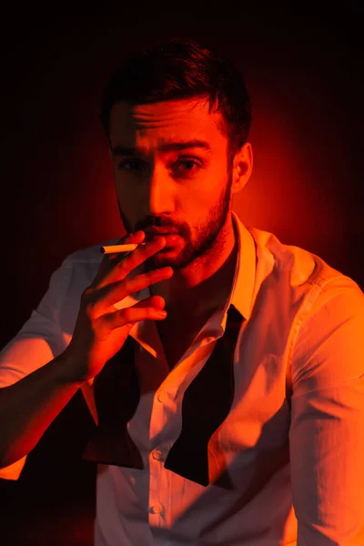 Бородатий чоловік у формальному носінні тримає сигарету на чорному тлі з червоним освітленням — стокове фото