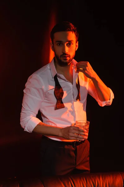 Elegante uomo barbuto con bicchiere di whisky in piedi su sfondo nero con illuminazione rossa — Foto stock