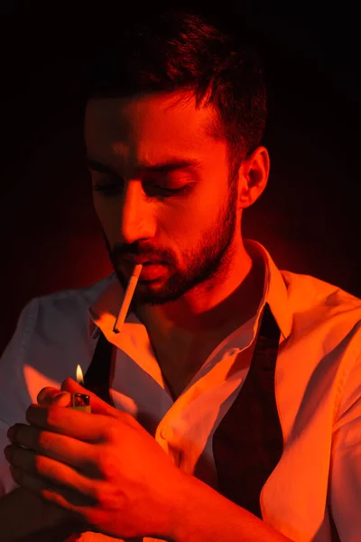 Hombre barbudo con cigarrillo sosteniendo encendedor en luz roja sobre fondo negro - foto de stock