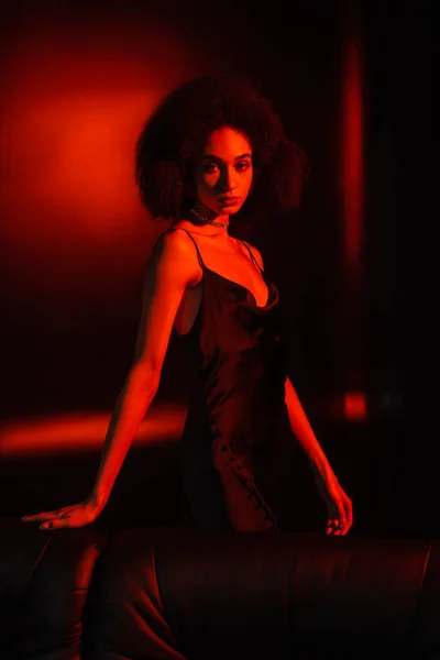African american woman in dress posing near couch on black background with red lighting — Stock Photo