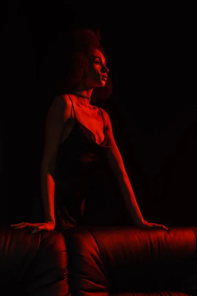 African american woman in dress posing near couch in red light isolated on black — Stock Photo