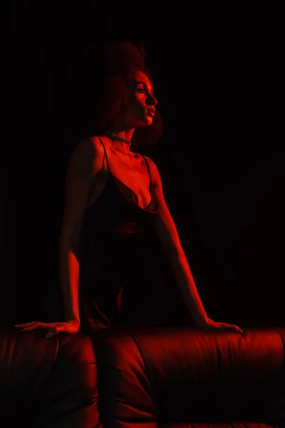 Mujer afroamericana en vestido posando cerca de sofá en luz roja aislado en negro - foto de stock