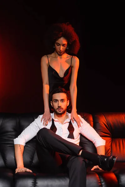 African american woman touching elegant boyfriend on couch on black background with red lighting — Stock Photo