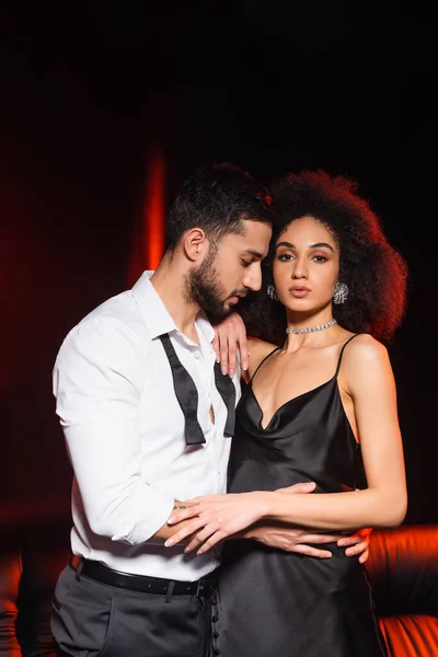 Man in formal wear hugging african american girlfriend in dress on black background with red lighting — Stock Photo