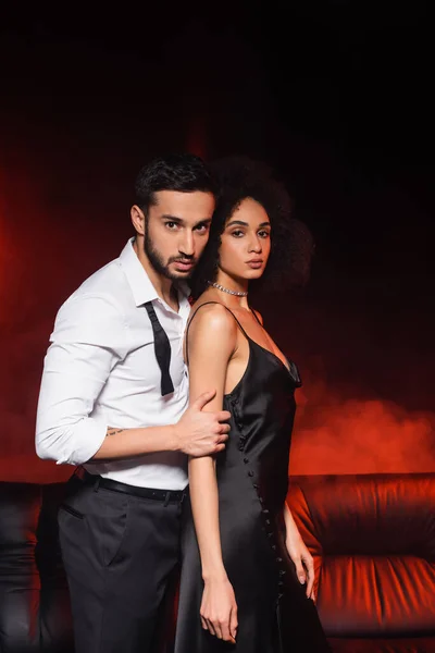 Man embracing african american girlfriend in dress on black background with red lighting and smoke — Stock Photo