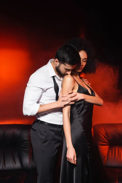 Bearded uomo baciare spalla di africano americano fidanzata su sfondo nero con rosso illuminazione e fumo — Foto stock
