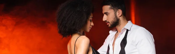 Side view of bearded man looking at african american girlfriend on black background with red lighting and smoke, banner — Stock Photo