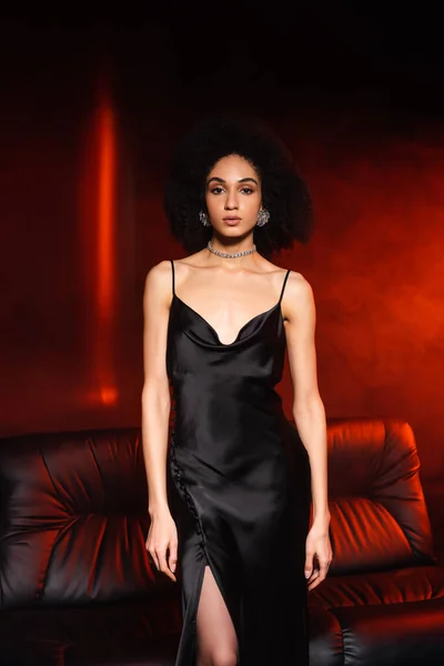 Elegante mujer afroamericana en vestido mirando a la cámara cerca del sofá sobre fondo negro con iluminación roja y humo - foto de stock