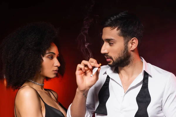 Man smoking cigarette near elegant african american girlfriend on black background with red lighting — Stock Photo