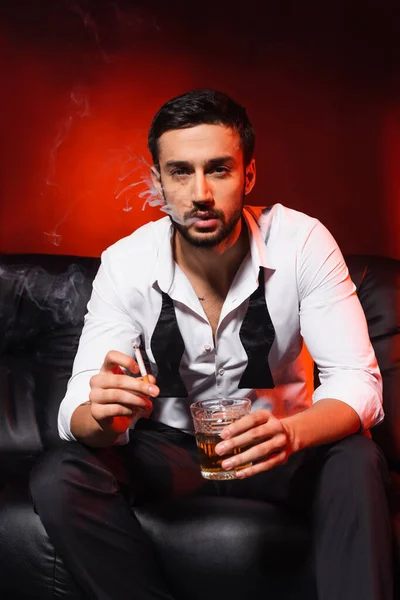 Elegant man smoking cigarette and holding glass of whiskey on couch on black background with red lighting — Stock Photo