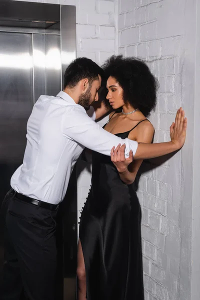 Hombre de pie cerca de la novia afroamericana en vestido cerca del ascensor - foto de stock