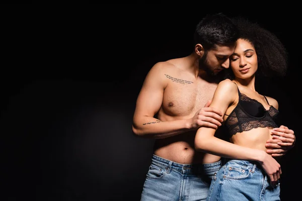 Hombre tatuado abrazando mujer afroamericana bonita en sujetador aislado en negro - foto de stock