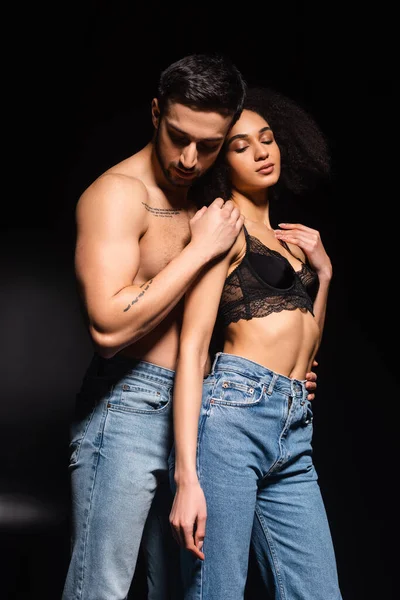 Hombre sexy abrazando a mujer afroamericana en lencería sobre fondo negro - foto de stock