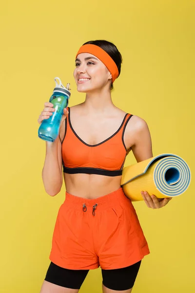 Glückliche Sportlerin mit Fitnessmatte trinkt Wasser aus Sportflasche isoliert auf gelb — Stockfoto