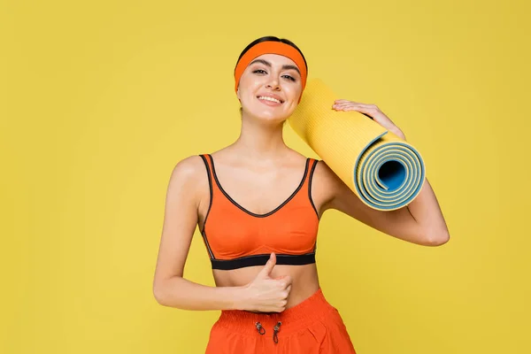 Cheerful sportswoman with fitness mat showing thumb up isolated on yellow - foto de stock