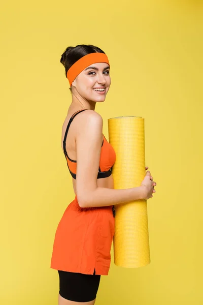 Smiling sportswoman with fitness mat looking at camera isolated on yellow — Stock Photo