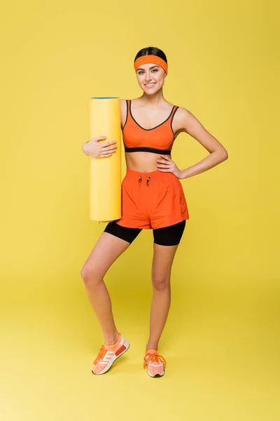 Visão de comprimento total de desportista alegre com tapete de fitness de pé com a mão no quadril no fundo amarelo — Fotografia de Stock