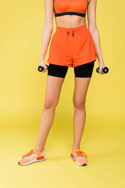 Обрезанный вид спортсменки в оранжевых шортах, стоящей с гантелями на желтом фоне — стоковое фото