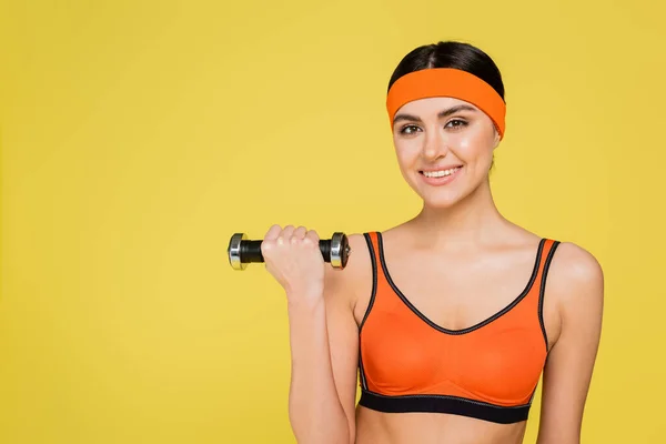Pretty sportswoman smiling at camera while training with dumbbell isolated on yellow — Stock Photo