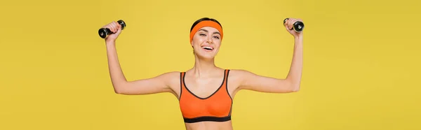 Весела жінка в помаранчевому спортивному бюстгальтері, що працює з гантелями ізольовано на жовтому, банер — стокове фото